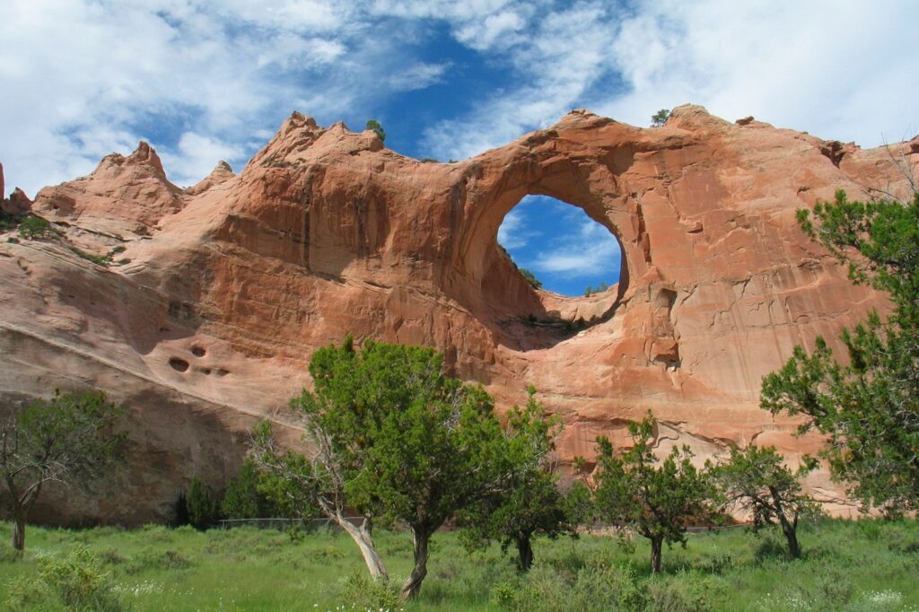 Large randstone rock formation with a round window-like hole in the center, surrounded by grass and trees.