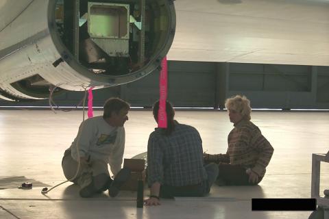 Three people sitting on the floor of an airplane hangar under a plane wing and engine that is being built/repaired